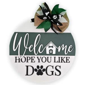 This charming round wooden door sign warmly greets visitors with the text, "Welcome Hope You Like Dogs." It showcases a small doghouse and playful paw prints, beautifully embellished with a burlap bow adorned with paw print ribbons.