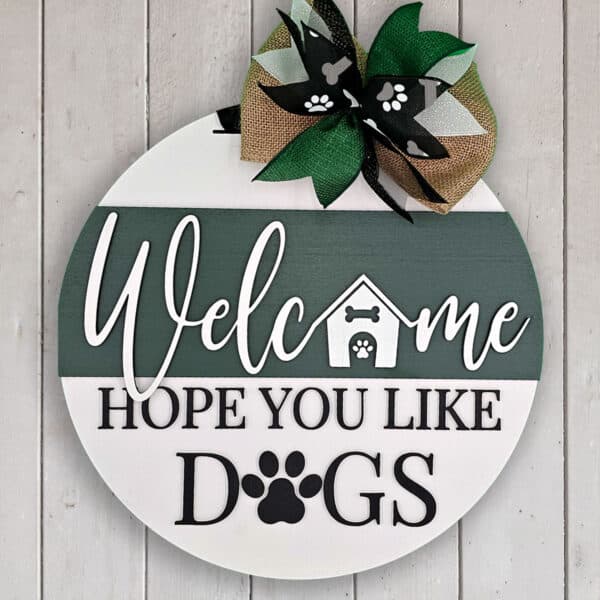 A round wooden welcome sign hangs against a white wall. The text, "Welcome, Hope You Like Dogs," is accented with paw prints and a dog house image. A patterned ribbon bow at the top adds charm, adorned with even more paw prints.