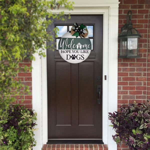 A dark brown front door with a glass panel at the top, set in a brick wall, surrounded by white trim. Above the door, a custom door sign reads, "Welcome, Hope You Like Dogs," with dog paw prints and a small house illustration. A garden lantern is on the right wall.