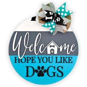 This charming round wooden door sign warmly greets visitors with the text, "Welcome Hope You Like Dogs." It showcases a small doghouse and playful paw prints, beautifully embellished with a burlap bow adorned with paw print ribbons.