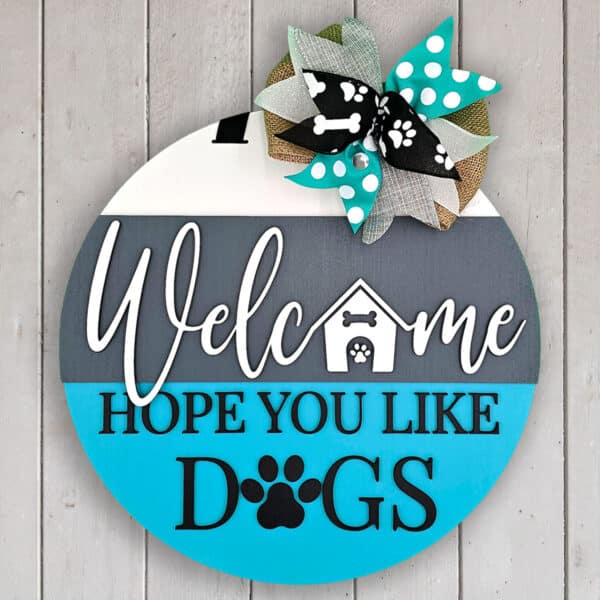 A round wooden welcome sign hangs against a white wall. The text, "Welcome, Hope You Like Dogs," is accented with paw prints and a dog house image. A patterned ribbon bow at the top adds charm, adorned with even more paw prints.