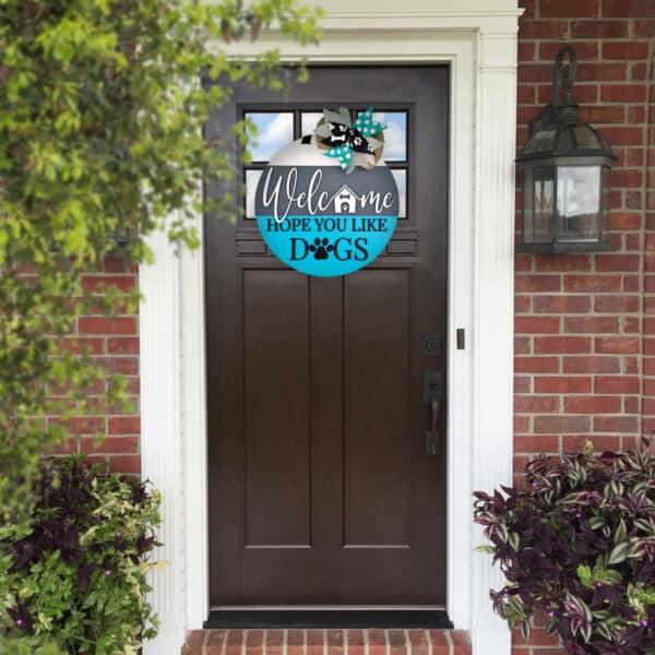 A dark brown front door with a glass panel at the top, set in a brick wall, surrounded by white trim. Above the door, a custom door sign reads, "Welcome, Hope You Like Dogs," with dog paw prints and a small house illustration. A garden lantern is on the right wall.