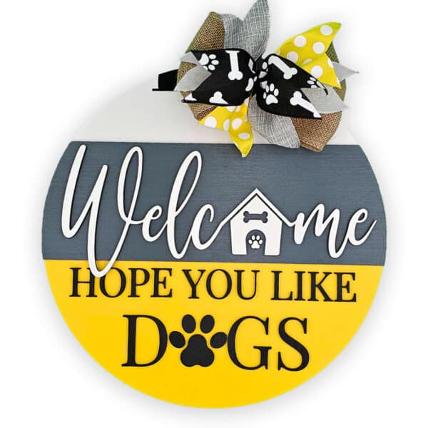 This charming round wooden door sign warmly greets visitors with the text, "Welcome Hope You Like Dogs." It showcases a small doghouse and playful paw prints, beautifully embellished with a burlap bow adorned with paw print ribbons.