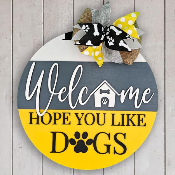 A round wooden welcome sign hangs against a white wall. The text, "Welcome, Hope You Like Dogs," is accented with paw prints and a dog house image. A patterned ribbon bow at the top adds charm, adorned with even more paw prints.