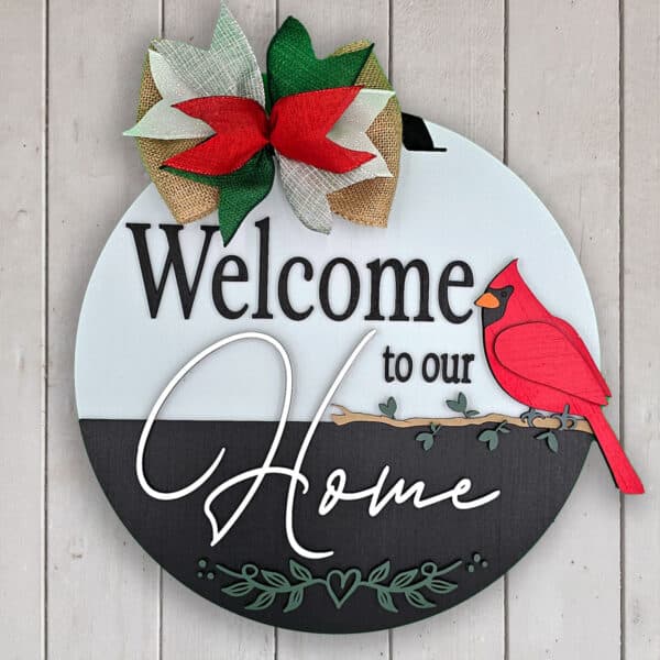 Wooden sign with "Welcome to our Home" in elegant letters. Features a red cardinal perched on a branch and a large bow with red, green, and burlap colors. The background is a light wood panel wall.