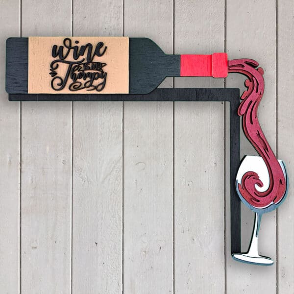 A decorative wooden wall art piece featuring a bottle pouring red wine into a glass. The bottle is inscribed with "Wine is my Therapy." The background consists of light wooden planks.